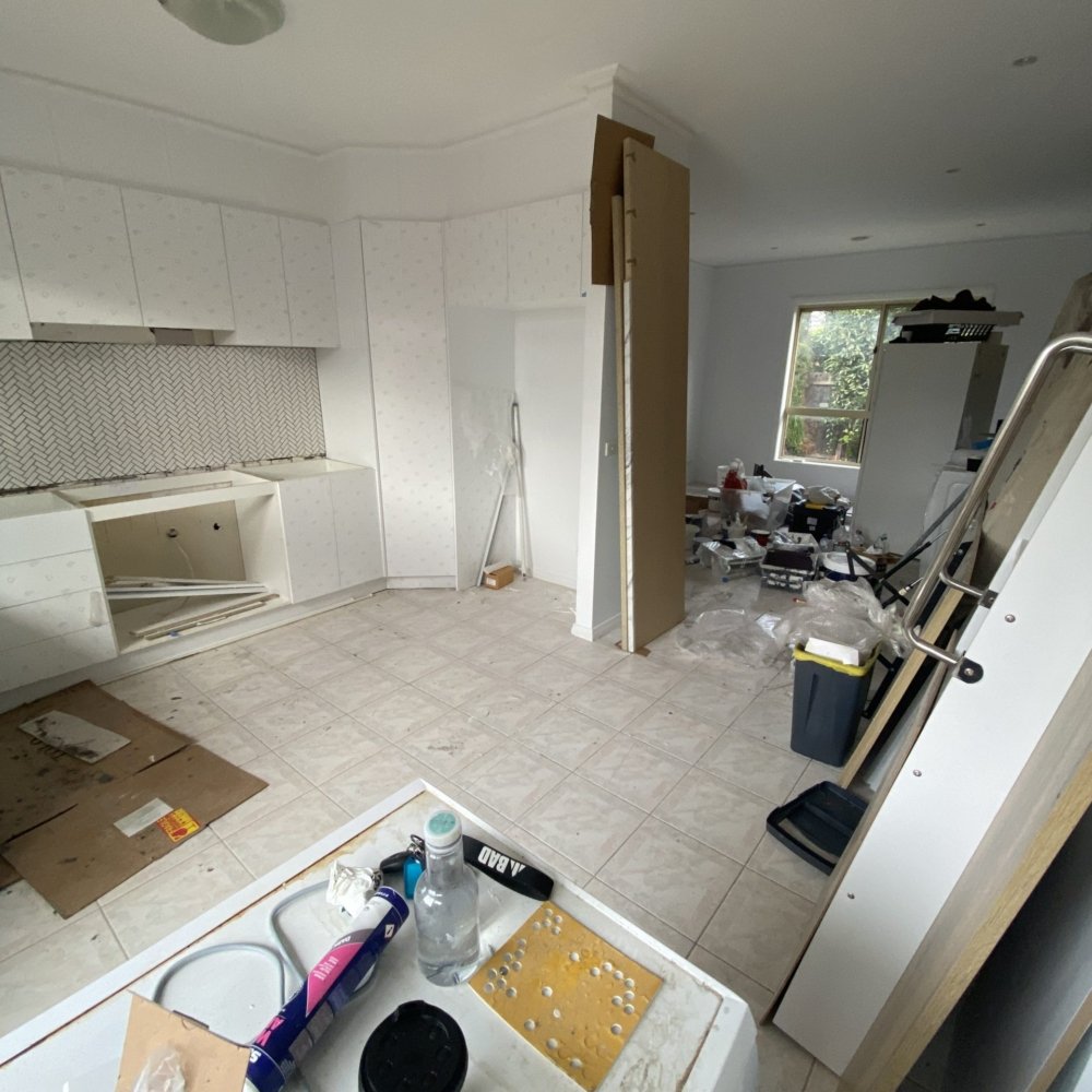 The kitchen during renovation shows bare walls, exposed plumbing, and tools scattered across the floor